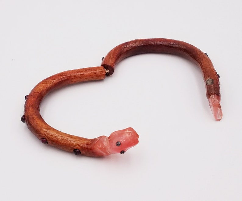 K&ouml;nnte beinhalten: Ein Holz-Armband mit einem rosa Schlangenkopf-Anh&auml;nger und roten Edelstein-Akzenten.