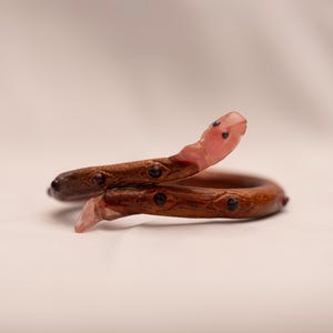May include: A pink and brown wooden snake bracelet with black accents. The snake is coiled and the head is raised.