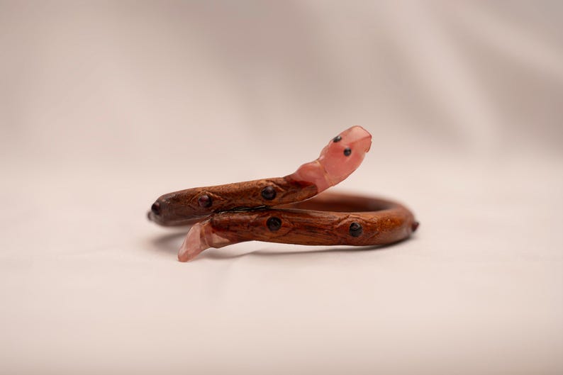 K&ouml;nnte beinhalten: Ein rosafarbenes und braunes Holzschlangenarmband mit schwarzen Akzenten. Die Schlange ist zusammengerollt und der Kopf ist erhoben.