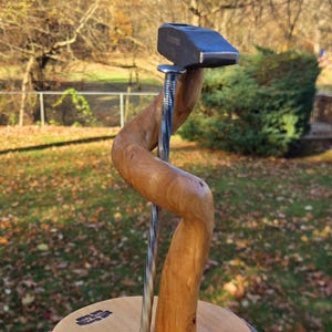 May include: A wooden sculpture with a hammer head, a twisted nail, and a curved wooden support. The sculpture is mounted on a round wooden base. The hammer head has the word "YOUNG" stamped on it.