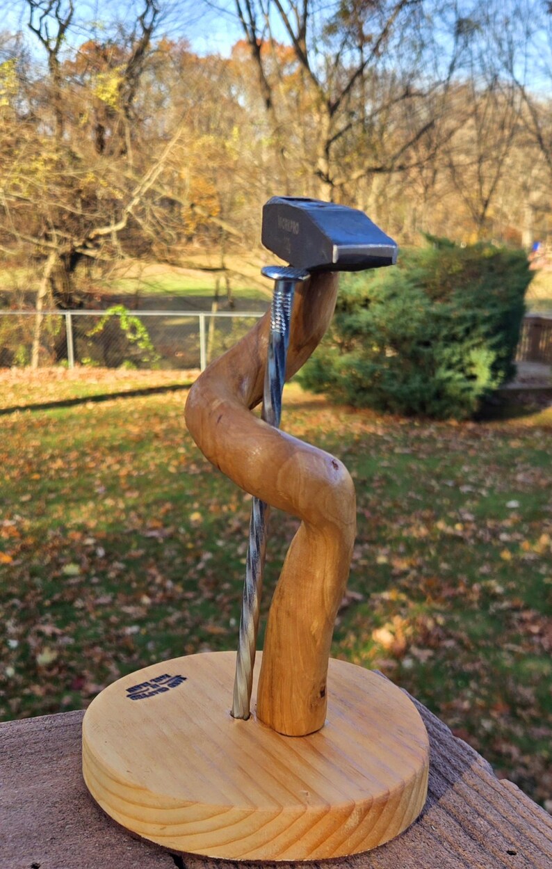 May include: A wooden sculpture with a hammer head, a twisted nail, and a curved wooden support. The sculpture is mounted on a round wooden base. The hammer head has the word "YOUNG" stamped on it.