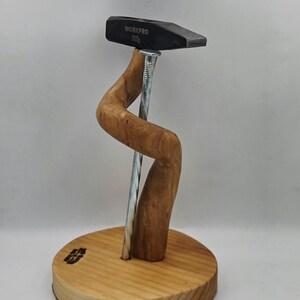 May include: A wooden sculpture of a hammer with "WORKPRO 200g" text, supported by a curved wooden element and a large nail. It's mounted on a round wooden base. The wood has a natural finish. The hammer is 200 grams.