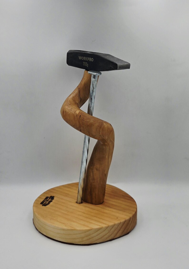 May include: A wooden sculpture of a hammer with "WORKPRO 200g" text, supported by a curved wooden element and a large nail. It's mounted on a round wooden base. The wood has a natural finish. The hammer is 200 grams.
