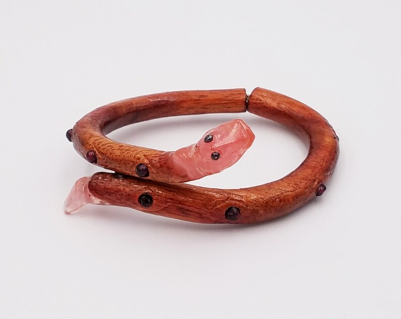 K&ouml;nnte beinhalten: Ein Holz-Armband mit einem rosafarbenen Schlangen-Anh&auml;nger. Das Armband ist mit kleinen, dunkelroten Edelsteinen verziert.