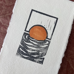 May include: A black and orange linocut print of a sunset over the ocean. The sun is a bright orange circle and the ocean is represented by black wavy lines.