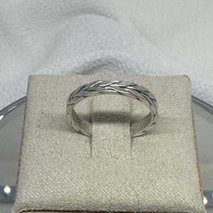 May include: A silver ring with a woven design. The ring is displayed on a beige square surface. The ring has a textured pattern around the band. The ring is a simple, elegant piece of jewelry.