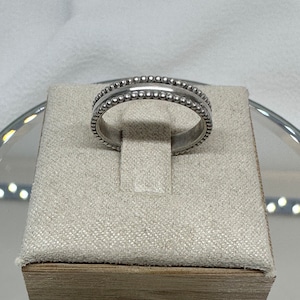May include: A silver ring with a beaded design. The ring features a band of small, raised beads along the top and bottom edges, with a smooth, recessed center. The ring is displayed on a beige, textured surface.