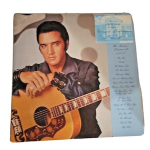 Puede incluir: Portada de álbum vintage de Elvis Presley. La imagen muestra a Elvis con una guitarra, vistiendo una camisa azul oscuro y una pulsera negra. El álbum tiene un fondo de color beige claro con un borde azul y texto. El álbum se titula "Command Performance".