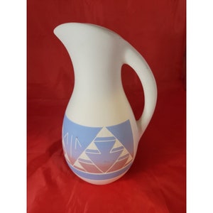 Può includere: Brocca in ceramica bianca con manico e beccuccio curvi. La brocca presenta un design geometrico nei toni del blu e del rosa nella parte inferiore. La brocca viene probabilmente utilizzata per servire bevande.