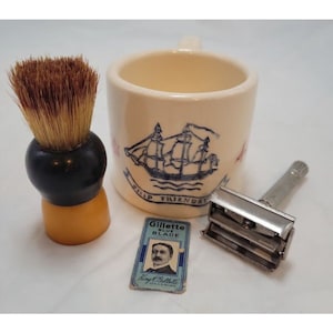 Pode incluir: Um conjunto de barbear vintage com um pincel de barbear com cabo preto e cerdas castanhas, uma caneca creme com uma ilustração de navio, uma navalha de segurança e um pacote de lâminas Gillette Blue.