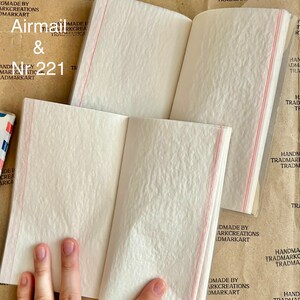 May include: A small, white, blank notebook with red lines on the spine. The notebook is open to reveal the blank pages inside. The cover of the notebook has the text "Airmail & N221" printed on it. The notebook is made by Trademark Creations.
