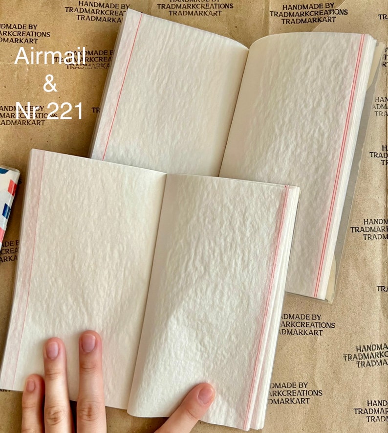 May include: A small, white, blank notebook with red lines on the spine. The notebook is open to reveal the blank pages inside. The cover of the notebook has the text "Airmail & N221" printed on it. The notebook is made by Trademark Creations.