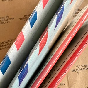 May include: Three red, white, and blue air mail envelopes stacked on top of each other. The envelopes are made of paper and have a red, white, and blue striped pattern. The envelopes are labeled "Air Mail" in black lettering.