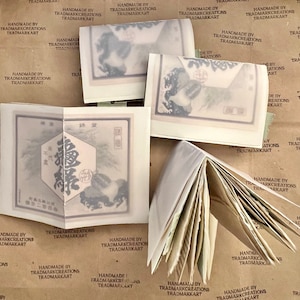 May include: A handmade book with a brown paper cover. The book is open to reveal pages with a light brown color. The book is surrounded by several small, folded pieces of paper with images of Japanese art. The paper has the text "Handmade by Trademarkcreations Trademarkart" printed on it.