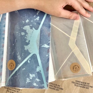 May include: Four handmade envelopes with geometric designs. The envelopes are made of translucent paper with a white background and a light blue, dark blue, or beige geometric pattern. Each envelope has a round brown sticker with the text "Handmade by Trademark Creations Trademarkart" on it.