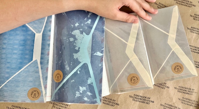 May include: Four handmade envelopes with geometric designs. The envelopes are made of translucent paper with a white background and a light blue, dark blue, or beige geometric pattern. Each envelope has a round brown sticker with the text "Handmade by Trademark Creations Trademarkart" on it.