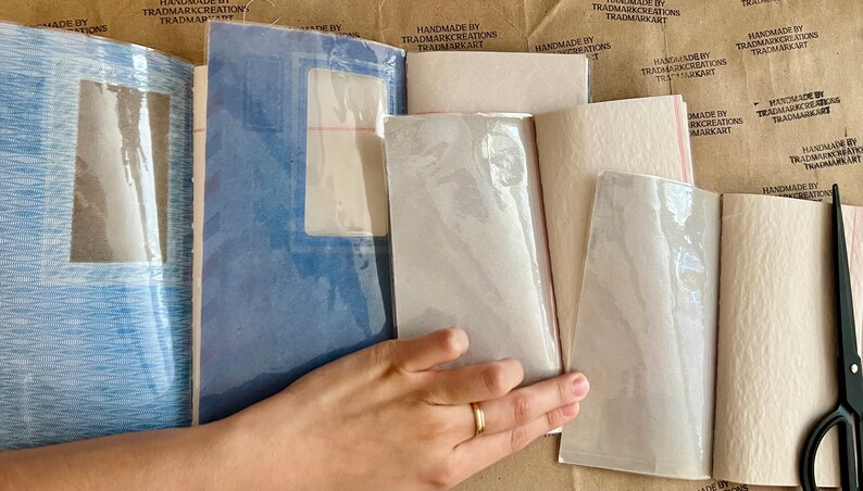 May include: A close-up of a hand holding a stack of handmade paper notebooks. The notebooks are covered in a clear plastic sheet and have a textured paper cover. The notebooks are labeled "Handmade by Trademark Creations Trademarkart".
