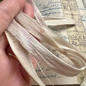 May include: Close-up of a white satin ribbon with a frayed edge. The ribbon is folded in half and is lying on a surface with handwritten text.