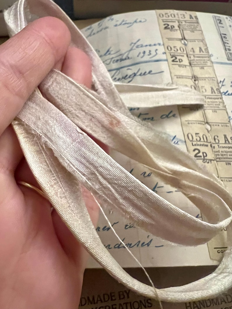 May include: Close-up of a white satin ribbon with a frayed edge. The ribbon is folded in half and is lying on a surface with handwritten text.