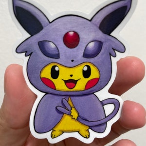 May include: A sticker of a yellow Pikachu wearing a purple and red Espeon costume. The sticker is on a hand.