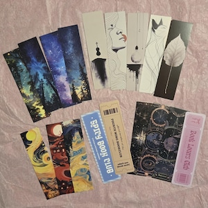 May include: A collection of illustrated bookmarks with various designs, including starry night scenes, forest landscapes, and abstract portraits. Some bookmarks feature text like "Spicy Book Club" and "Book Lovers Club."