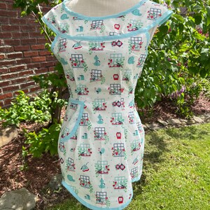 May include: A white apron with a blue trim and pocket, featuring a repeating pattern of garden scenes. The apron is displayed on a mannequin in a garden setting, showcasing its design and style.