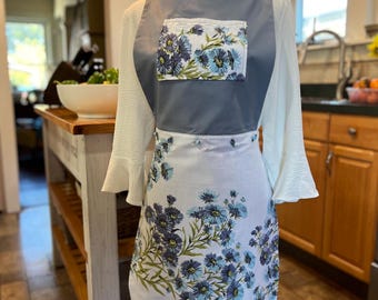 May include: A white and blue floral apron with a gray top and pocket. The apron features a ruffle at the bottom and is displayed on a mannequin. The kitchen apron has a floral pattern with blue and green accents.