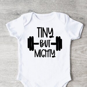 May include: White baby onesie with the text "TINY BUT MIGHTY" in black, featuring a barbell graphic. The onesie is short-sleeved and has a simple, classic design, perfect for a newborn or infant.