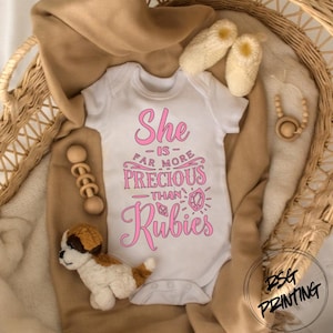 May include: White baby onesie with pink text that reads "She is far more precious than Rubies." The onesie is surrounded by a tan blanket, a stuffed puppy, wooden beads, and a pair of cream-colored baby booties.