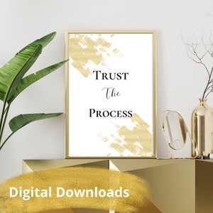 May include: A white printable poster with gold brushstrokes and the text "TRUST The PROCESS" in black. The poster is framed in a gold frame and is displayed on a gold shelf.