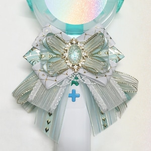 May include: A light blue and white light stick with a round, iridescent sticker that says "PURPLE BYUL". The light stick has a white bow with gold accents and a light blue ribbon.