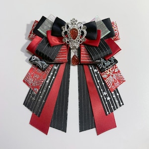 May include: A decorative bow brooch featuring red, black, and silver ribbons. The brooch is adorned with a central jewel-encrusted ornament and a dangling red gem. The design incorporates various patterns and textures, creating a layered effect.