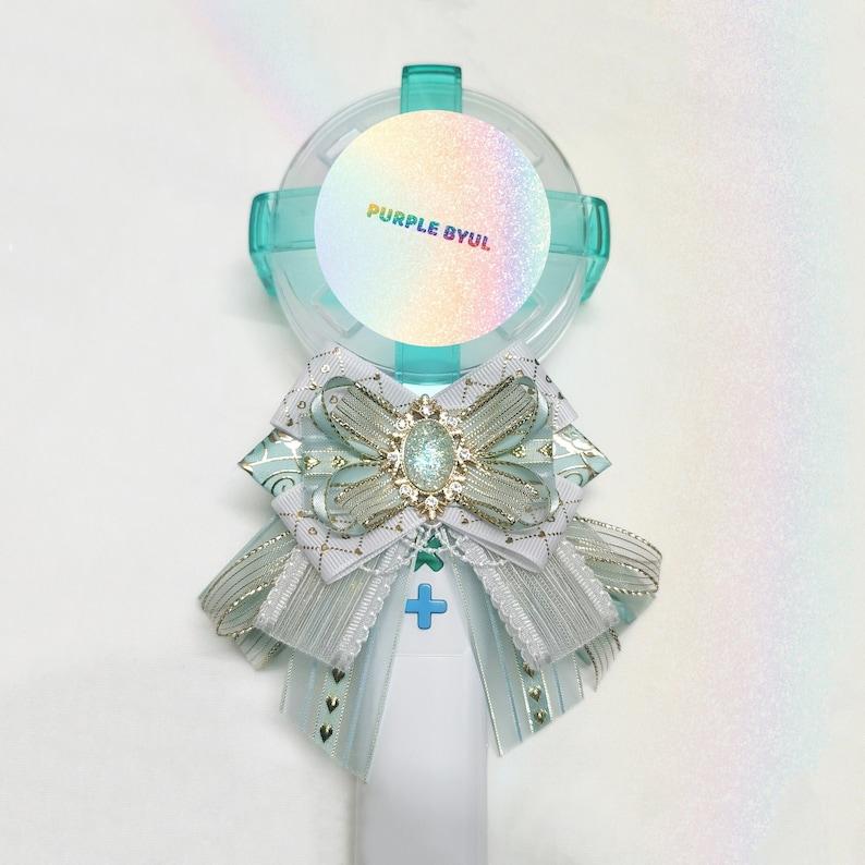 May include: A light blue and white light stick with a round, iridescent sticker that says "PURPLE BYUL". The light stick has a white bow with gold accents and a light blue ribbon.