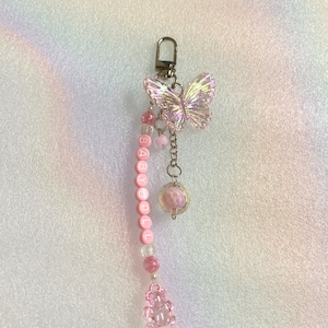 May include: A pink beaded keyring featuring a butterfly charm, the word "GABYDOLL" in white letters, a pink bear charm, and a clear, round charm with a pink flower inside.