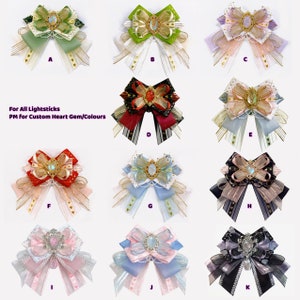 May include: Assortment of 11 handmade bows with various colors and patterns, including green, red, blue, pink, and black. Each bow features a decorative heart-shaped gem in the center. The text "For All Lightsticks PM for Custom Heart Gem/Colours" is displayed below the bows.