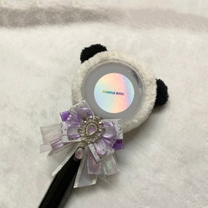 May include: A white and black panda-shaped light stick with a purple and iridescent sticker that says "PURPLE BYUL". The light stick has a purple and white bow with a rhinestone embellishment.
