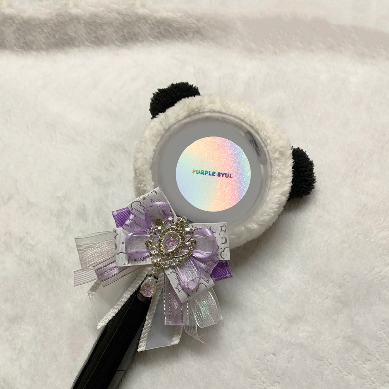May include: A white and black panda-shaped light stick with a purple and iridescent sticker that says "PURPLE BYUL". The light stick has a purple and white bow with a rhinestone embellishment.