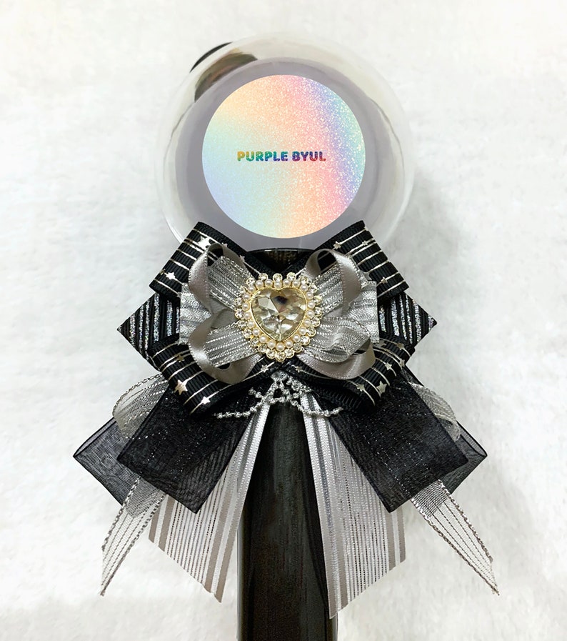 May include: A black and silver bow with a heart-shaped rhinestone embellishment sits atop a black and silver striped stick. The stick is topped with a clear plastic sphere with a rainbow glitter background and the text "PURPLE BYUL" in rainbow letters.