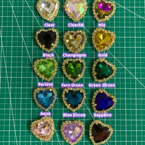 May include: Assortment of heart-shaped gems in various colors, including clear, black, gold, green, blue, pink, purple, and brown. The gems are set in gold-toned settings and are arranged in rows on a green grid background. The text "Heart Gems" is at the top of the image. The text "Siam, Rose, Peach, Black Diamond & Smoked Topaz also available (PM)" is at the bottom of the image.