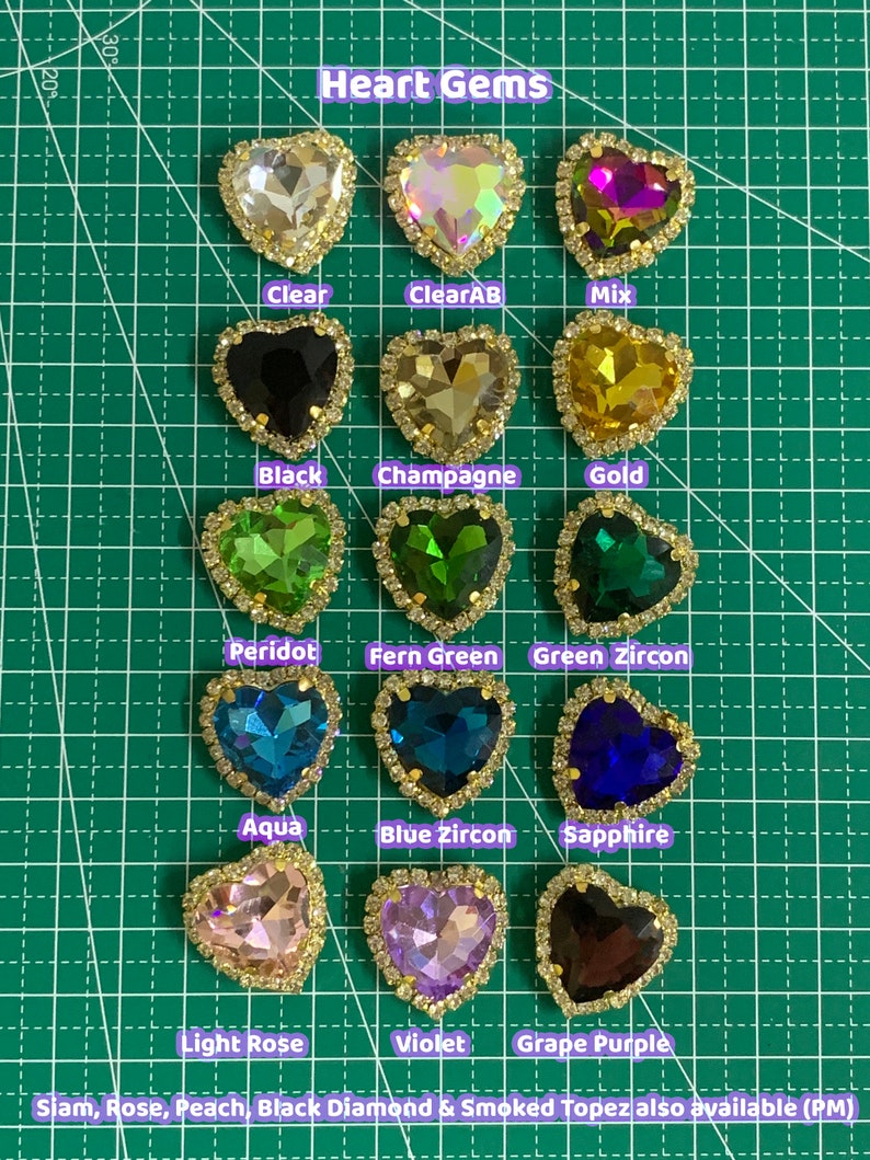 May include: Assortment of heart-shaped gems in various colors, including clear, black, gold, green, blue, pink, purple, and brown. The gems are set in gold-toned settings and are arranged in rows on a green grid background. The text "Heart Gems" is at the top of the image. The text "Siam, Rose, Peach, Black Diamond & Smoked Topaz also available (PM)" is at the bottom of the image.