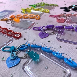 May include: A collection of colorful keychain charms with clear plastic frames and chain links. The charms feature a heart-shaped charm with the text "Pizza Piano" and a gummy bear charm.