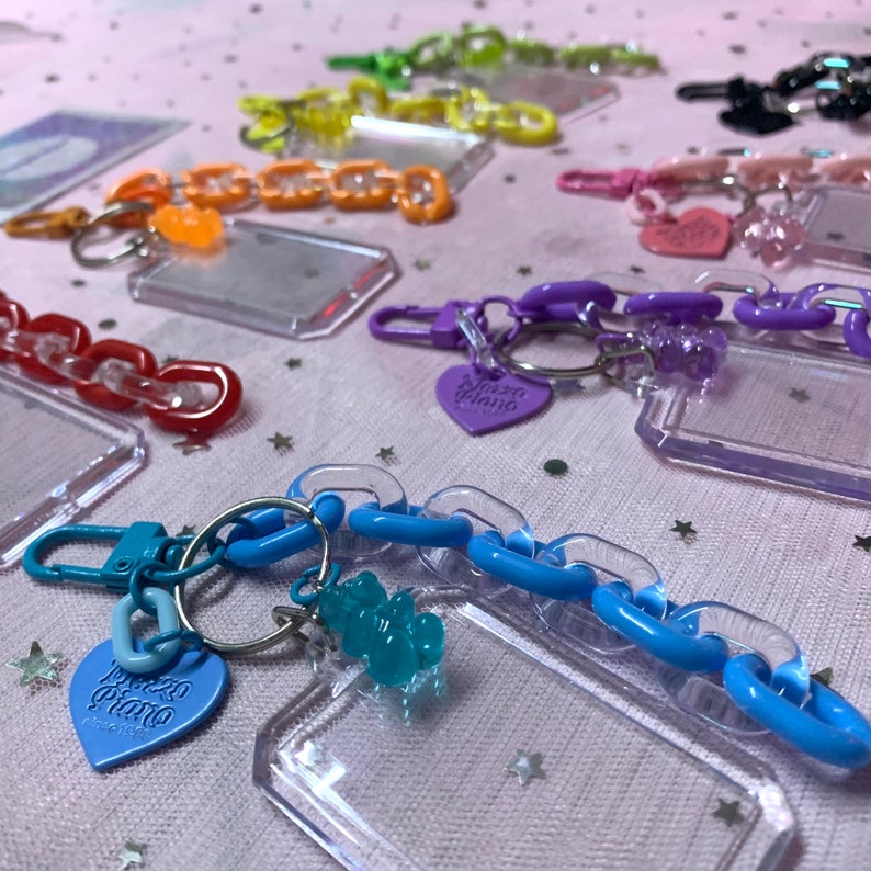 May include: A collection of colorful keychain charms with clear plastic frames and chain links. The charms feature a heart-shaped charm with the text "Pizza Piano" and a gummy bear charm.