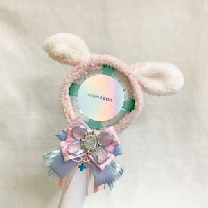 May include: A white and pink light-up fan with a fluffy bunny ear headband. The fan has a clear plastic cover with a holographic sticker that says "PURPLE BYUL". A pink and blue bow with a rhinestone embellishment is attached to the bottom of the fan.