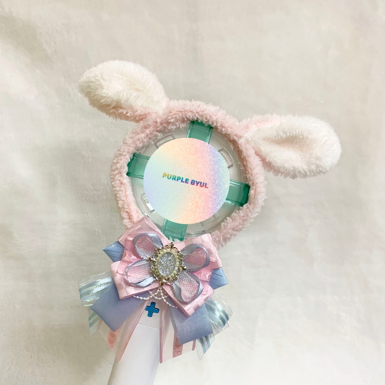 May include: A white and pink light-up fan with a fluffy bunny ear headband. The fan has a clear plastic cover with a holographic sticker that says "PURPLE BYUL". A pink and blue bow with a rhinestone embellishment is attached to the bottom of the fan.