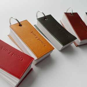 May include: Five small leather-bound notebooks with metal rings. Each notebook has the words "Good Luck" embossed on the cover. The notebooks are in different colours: red, orange, green, brown, and black.