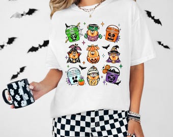 Könnte beinhalten: Weißes T-Shirt mit Halloween-Motiv mit verschiedenen Comicfiguren in Kostümen und Eimern. Die Figuren sind unter anderem ein Kürbis, ein Geist, eine Hexe und eine Mumie. Das Shirt hat ein schwarz-weißes Karomuster.