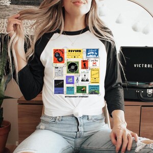 May include: White and black raglan sleeve t-shirt with a colorful graphic of vintage record album covers. The shirt has the words "Bittersweet Symphony" printed on it. A black Victrola record player is in the background.