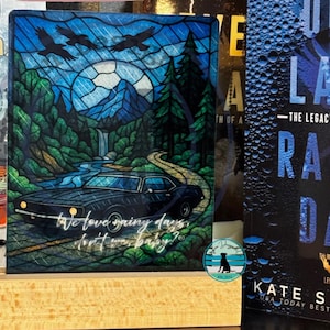 Puede incluir: Panel de vidrio decorativo con un paisaje estilizado con un coche negro en una carretera, montañas, árboles y tres cuervos contra un cielo azul. El texto "We love rainy days, don't we baby?" está escrito en el panel.