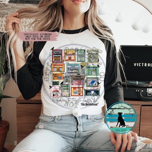 Könnte beinhalten: Weißes und schwarzes T-Shirt im Raglan-Stil mit einer farbenfrohen Grafik von Briefmarken mit verschiedenen Shop-Illustrationen. Das Shirt enthält den Text "Welcome to Triple Falls, NC" und "The Ravenhood". Im Hintergrund ist ein schwarzer Victrola-Plattenspieler zu sehen.