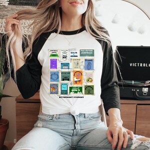 Peut inclure: Un t-shirt de baseball blanc et noir avec un graphique coloré d'un collage d'images. Le graphique comprend un terrain de football, un tourne-disque, une planche à roulettes, un horizon urbain et d'autres images. Le texte "BITTERSWEET SYMPHONY" est imprimé sous le graphique.